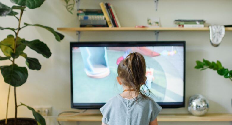 Image shows the back of a young girl, She is sat in a lounge watching TV.