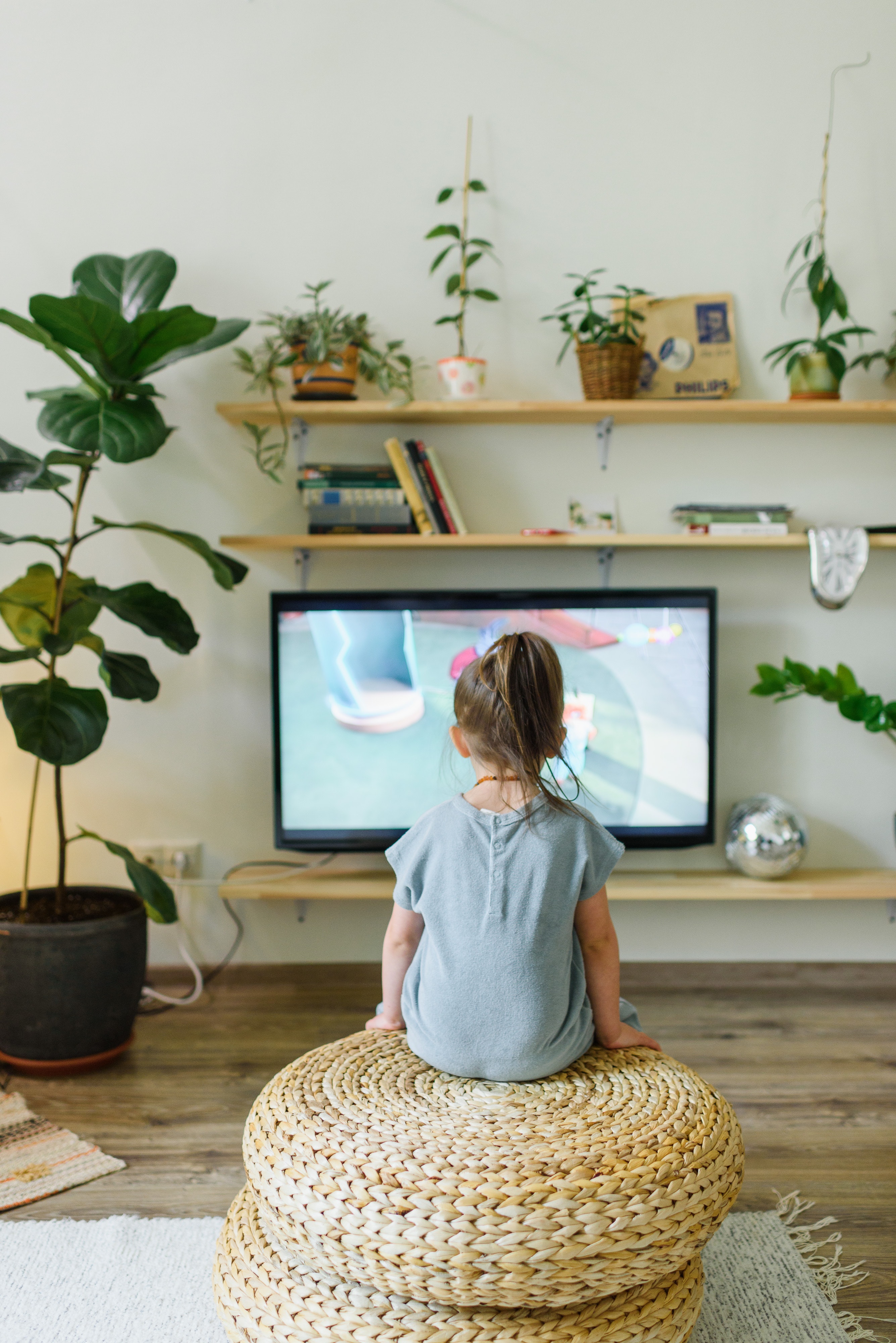 Image shows the back of a young girl, She is sat in a lounge watching TV.