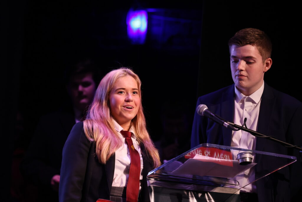 Image shows a young female at a lectern talking, with a young man at her side, 